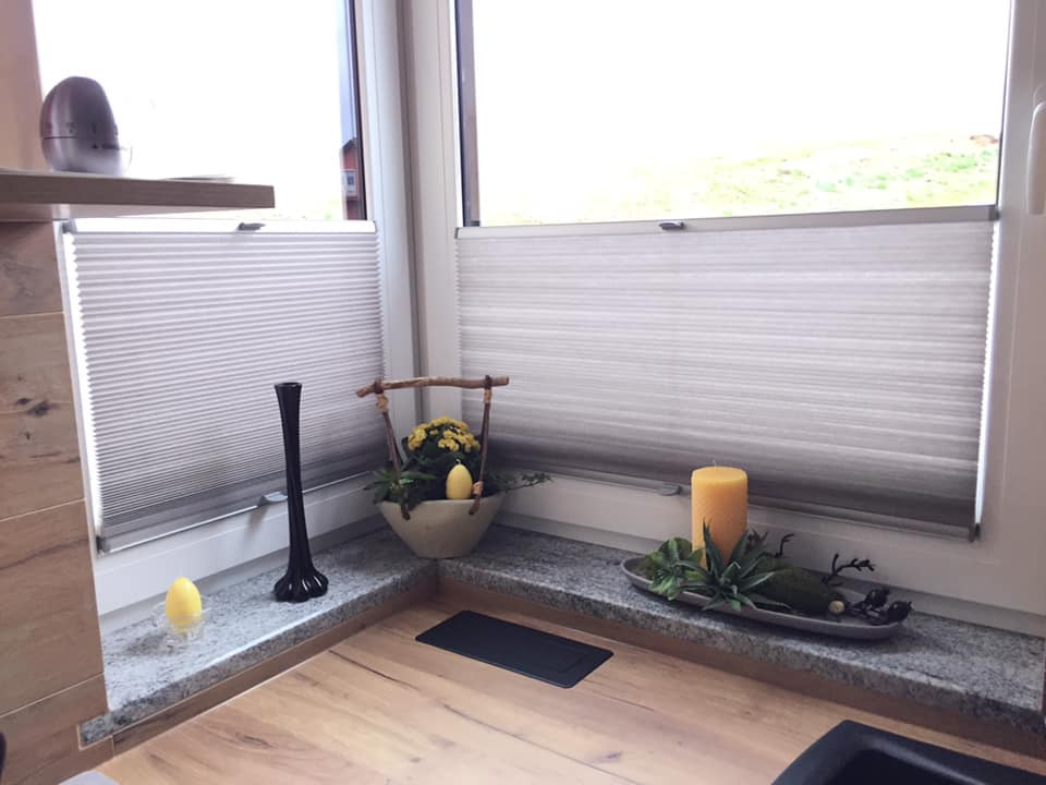 Dwa okna z plisowanymi osłonami okiennymi w kolorze szarym, dekorowane elementami wielkanocnymi, w tym żółtą świecą i koszyczkiem z kwiatami.