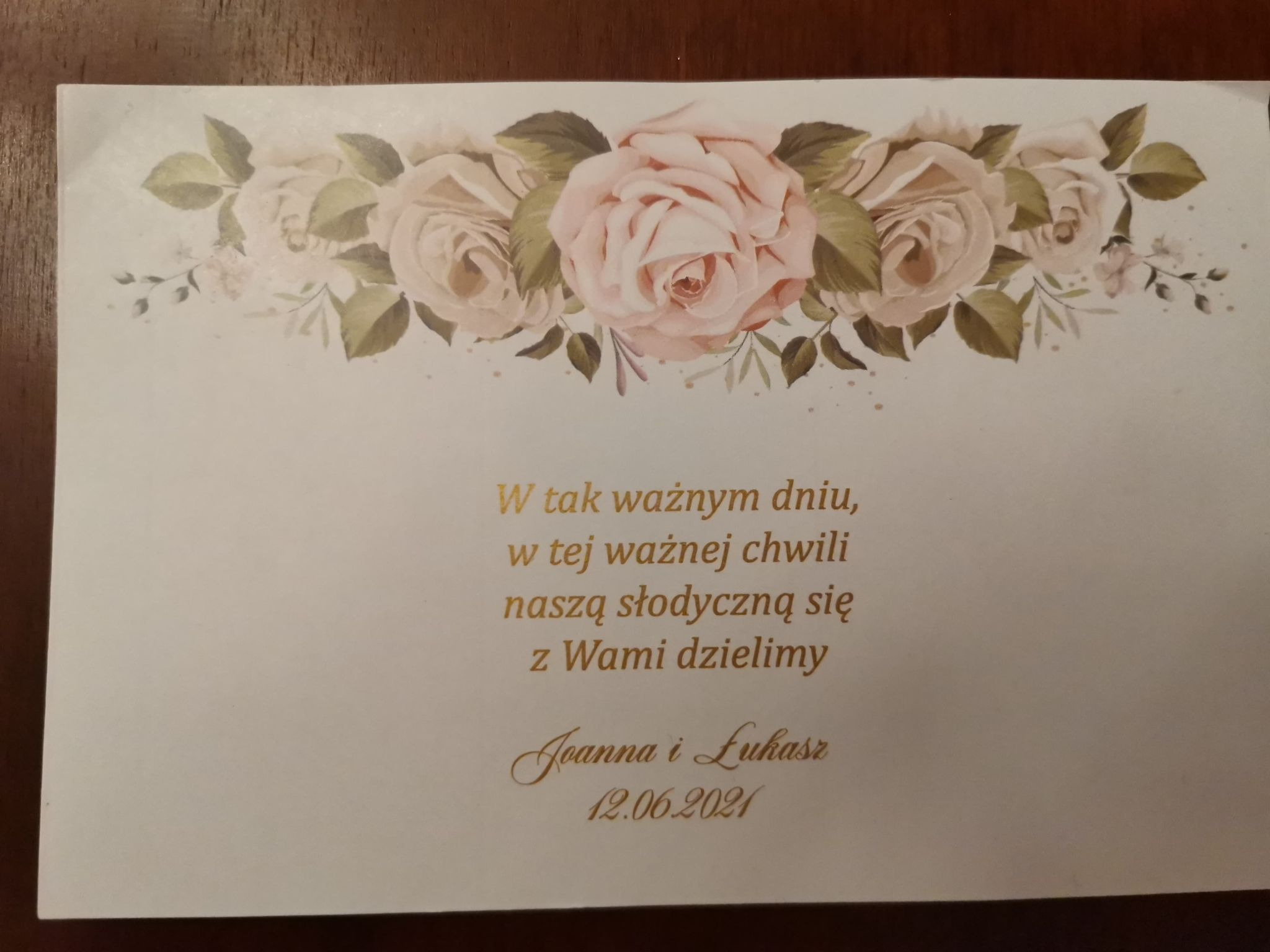 Eleganckie zaproszenie ślubne z motywem róż, delikatnym złoceniem tekstu i datą 12.06.2021, leżące na drewnianym stole.