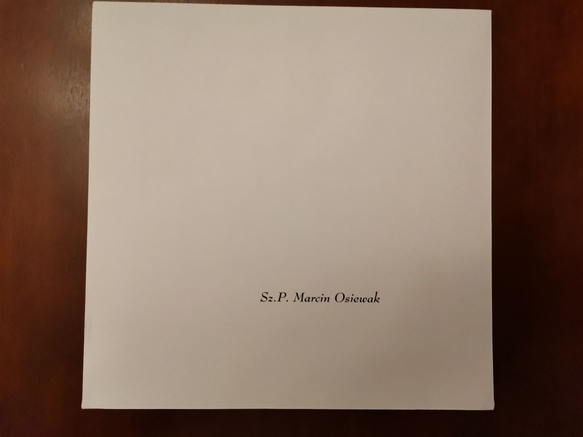 Biała kartka z czarnym tekstem 'Sz.P. Marcin Osiewak' leżąca na ciemnobrązowym drewnianym stole.