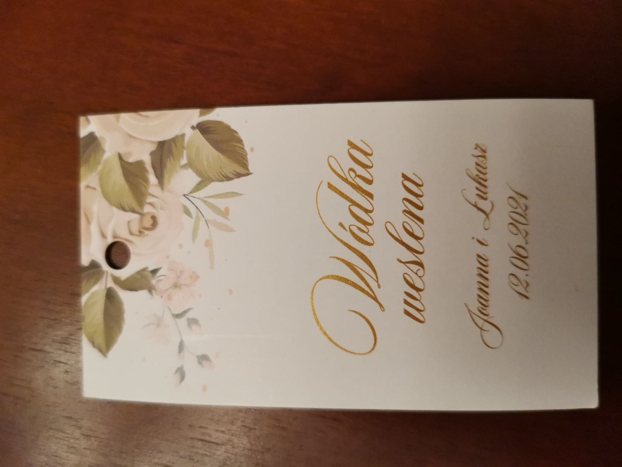 Personalizowana zawieszka na alkohol weselny z motywem kwiatowym, imionami Joanny i Łukasza oraz datą 12.06.2021, wykonana z białego papieru ze złoconym napisem.
