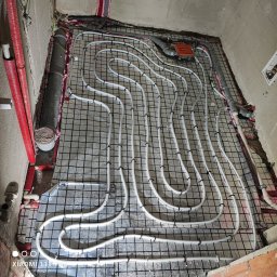 Tech House Tomasz Hauser - Ułożona instalacja ogrzewania podłogowego na siatce, widoczne białe rurki i czerwone rury przy ścianie. Etap prac przed wylaniem posadzki.