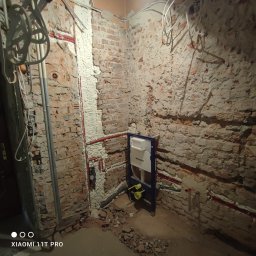 Tech House Tomasz Hauser - Montaż stelaża WC Geberit na surowej, ceglanej ścianie z instalacją wodną w pianie montażowej, Kraków. Widoczne przewody elektryczne.