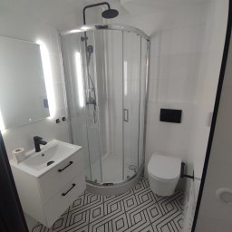 Tech House Tomasz Hauser - Nowoczesna łazienka po remoncie: białe płytki, kabina prysznicowa z czarnym deszczownicą, umywalka z szafką i czarną armaturą, toaleta oraz wzorzysta podłoga.