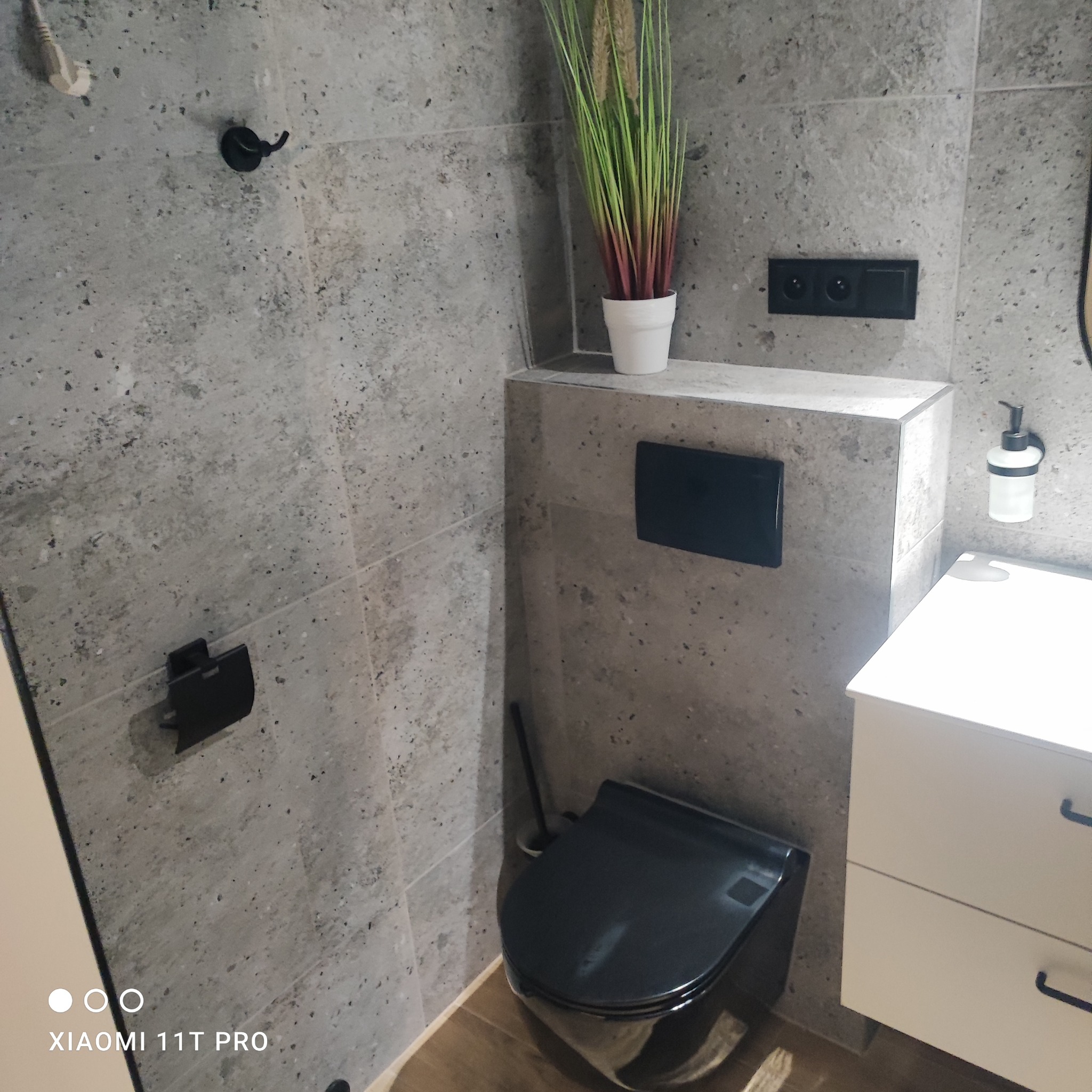 Nowoczesna toaleta w szarościach z czarnymi akcentami: wisząca miska WC, armatura, dozownik mydła. Uwagę zwraca minimalistyczny design i roślinny akcent dekoracyjny.