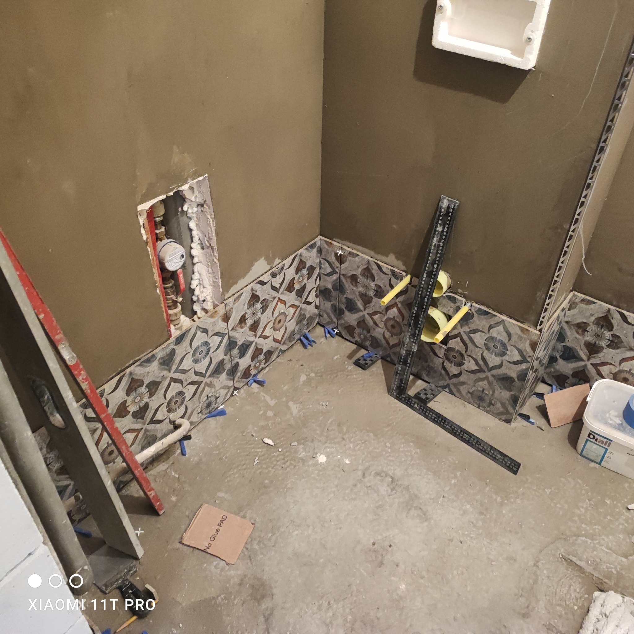 Układanie dekoracyjnych płytek ściennych w łazience. Widoczne narzędzia glazurnicze, poziomica i fragment instalacji wodnej w ścianie. Etap prac wykończeniowych.