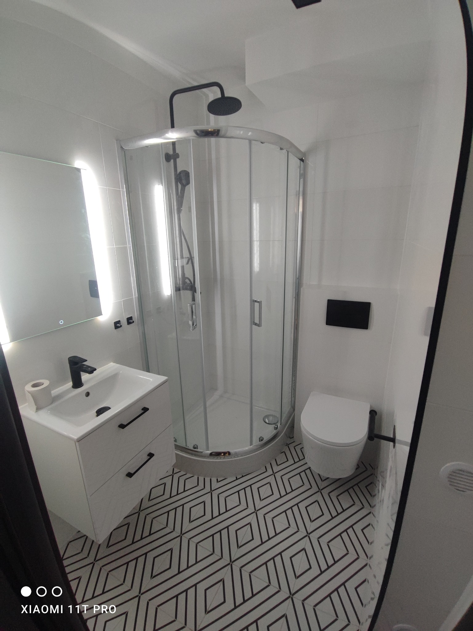 Nowoczesna łazienka po remoncie: białe płytki, kabina prysznicowa z czarnym deszczownicą, umywalka z szafką i czarną armaturą, toaleta oraz wzorzysta podłoga.