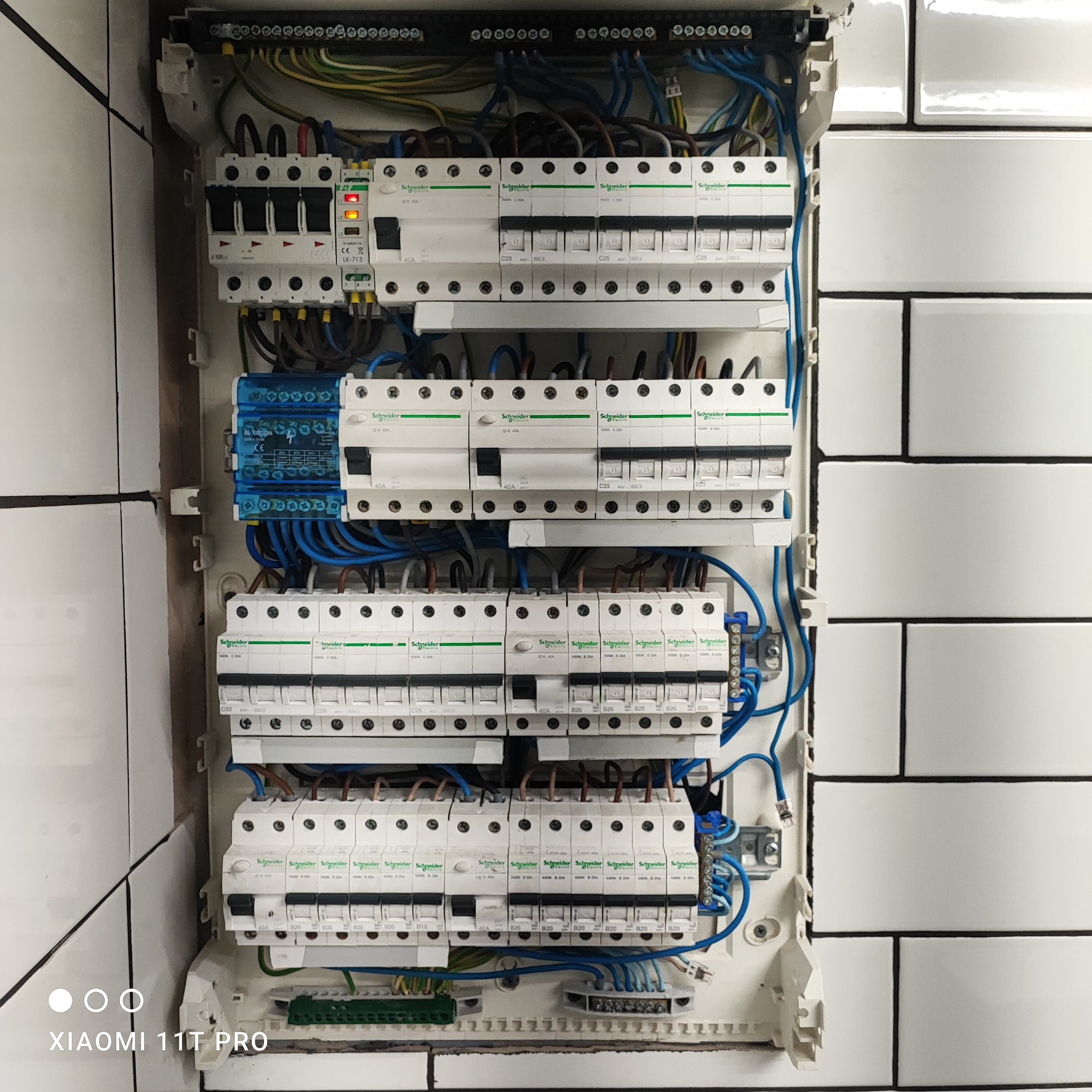 Rozdzielnica elektryczna Schneider z licznymi bezpiecznikami i okablowaniem, zamontowana na ścianie z białych płytek w stylu retro.