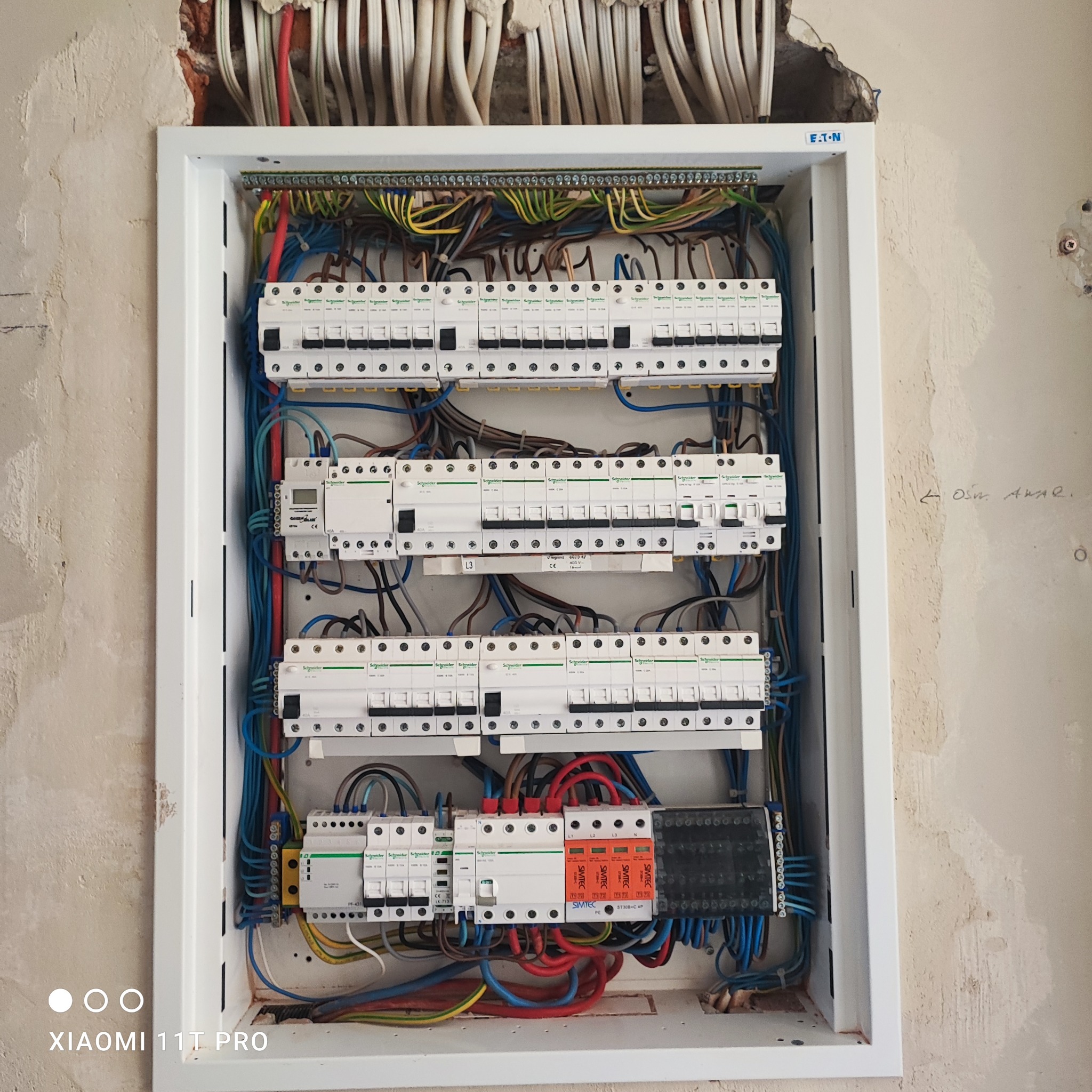 Rozdzielnica elektryczna z zainstalowanymi bezpiecznikami i przewodami w białej obudowie, widoczne kable w ścianie. Instalacja w trakcie prac wykończeniowych.