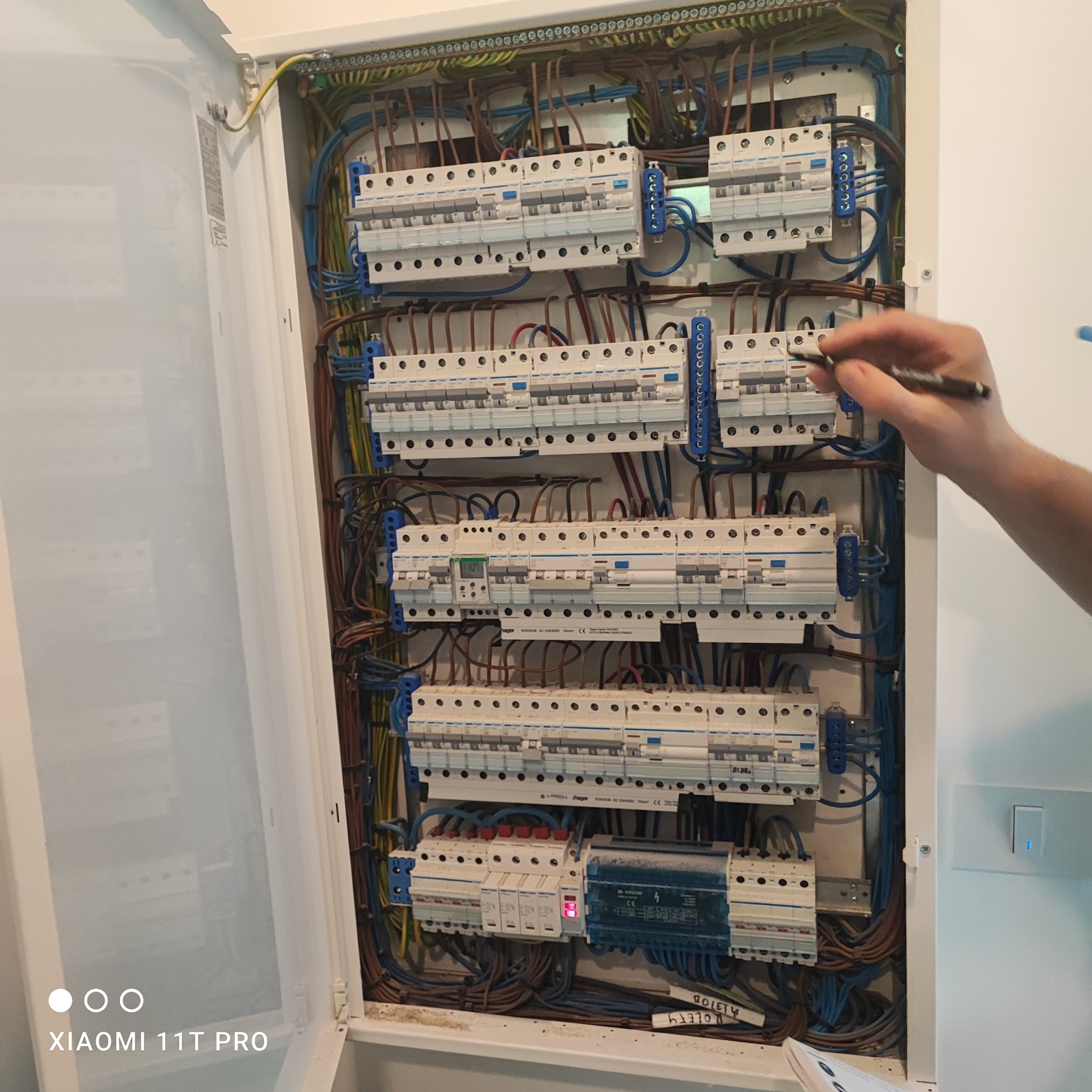 Otwarta skrzynka elektryczna z bezpiecznikami i przewodami. Widoczna dłoń z długopisem wskazująca na elementy instalacji. Realizacja instalacji elektrycznej.