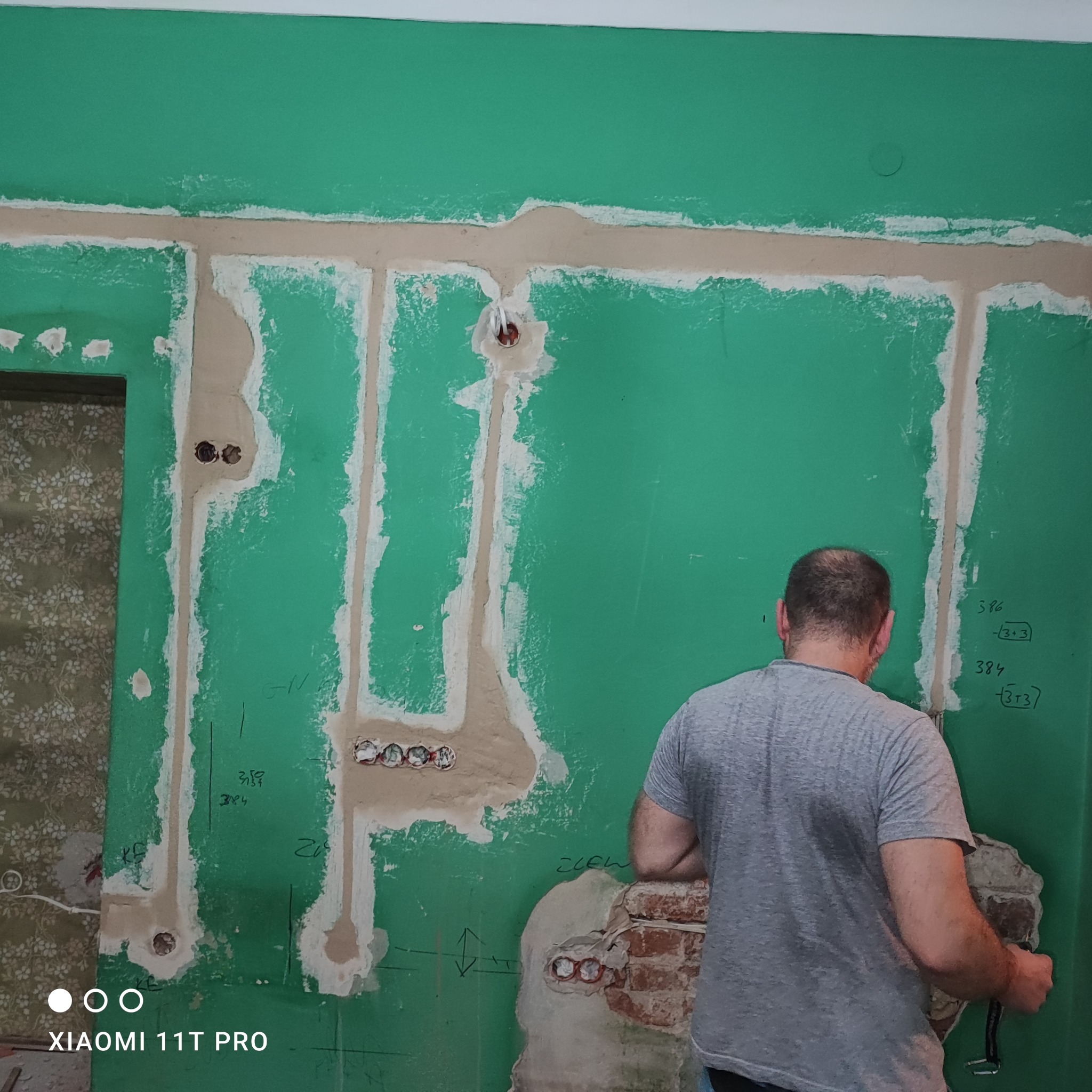 Bruzdowanie ścian pod instalację elektryczną. Widoczne kable w ścianie, przygotowane otwory pod gniazdka. Fachowiec w trakcie pracy. Zielona ściana z oznaczeniami.