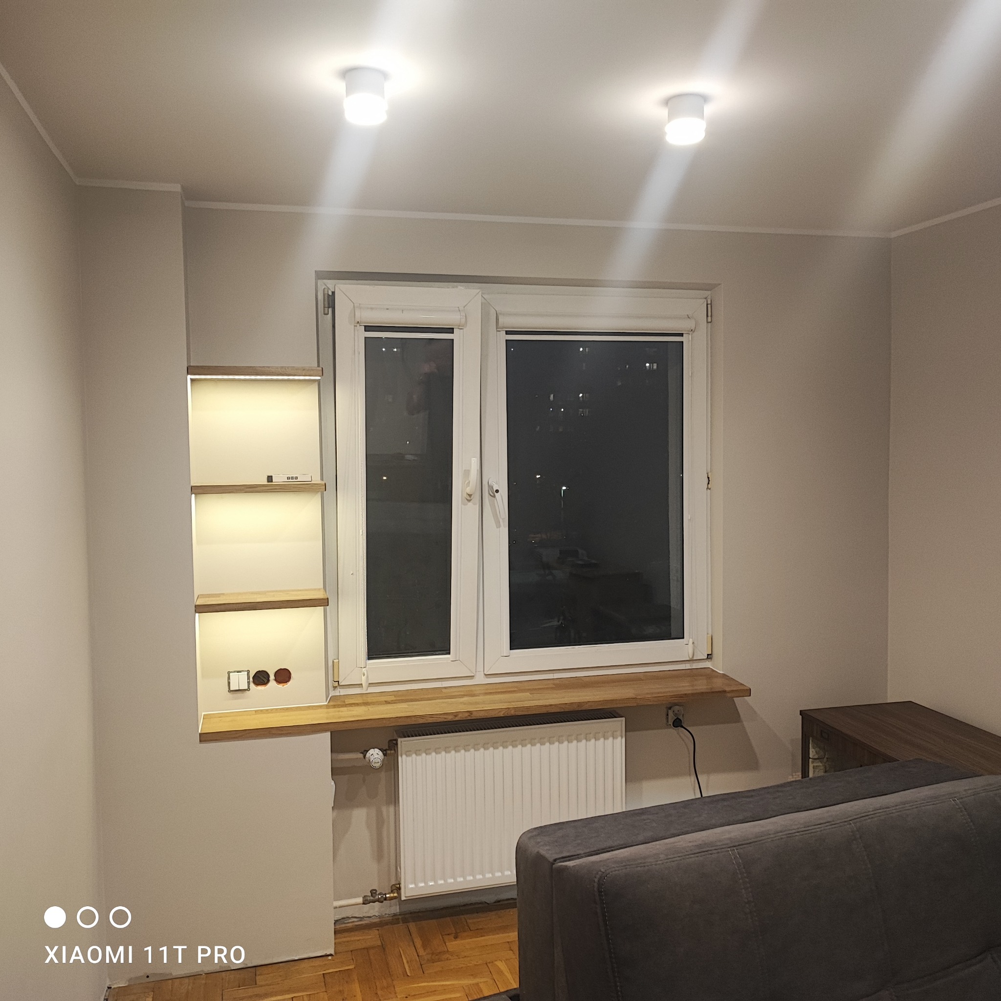 Białe okno z drewnianym parapetem i grzejnikiem. Obok wnęka z oświetlonymi półkami, widoczne gniazdka elektryczne. Nowoczesne wykończenie wnętrza.