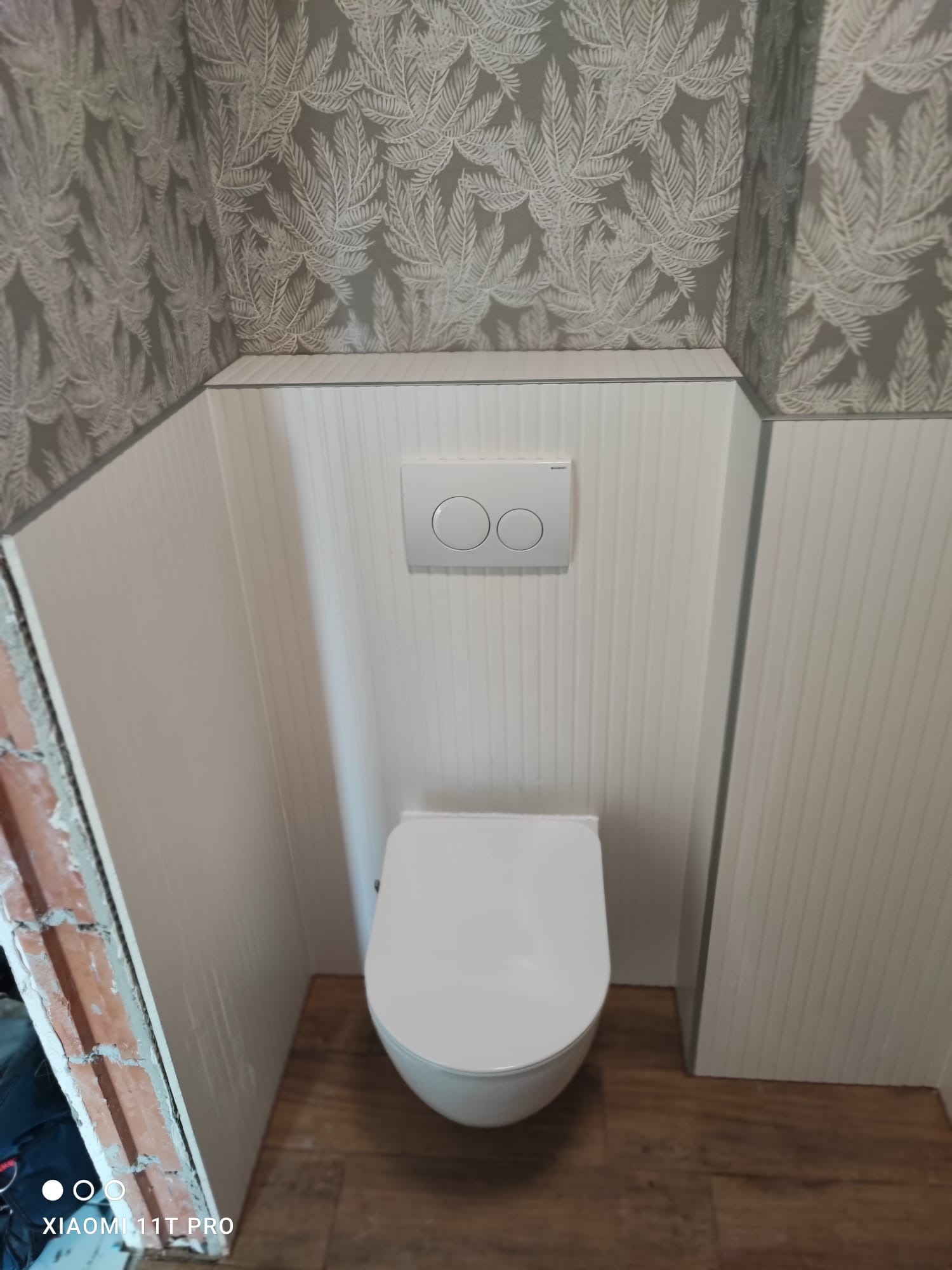 Wisząca toaleta z białym panelem spłukującym na tle ściany z tapetą w liście. Widoczne wykończenie podłogi i fragment ściany w trakcie prac wykończeniowych.