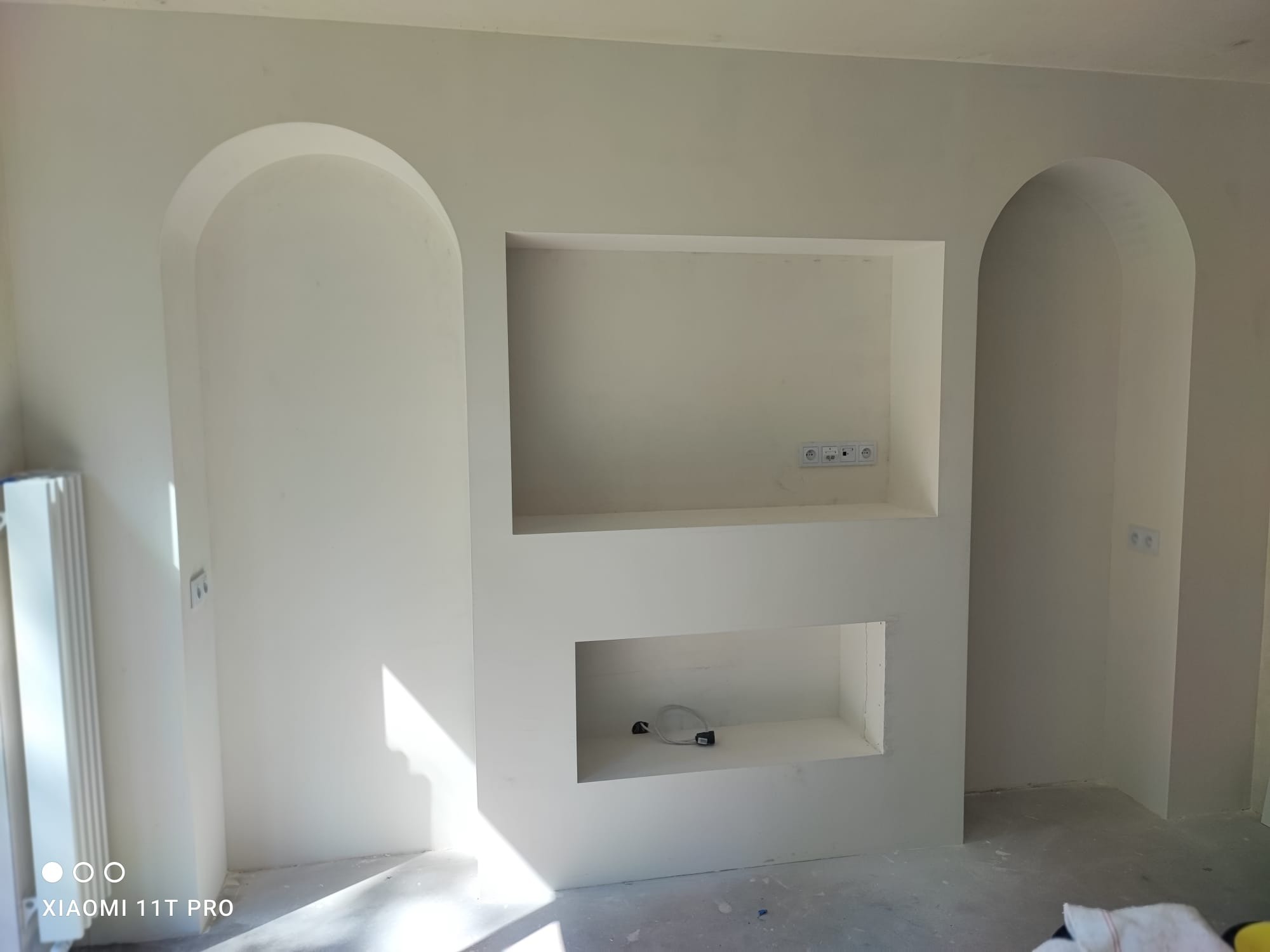 Białe ściany z wnękami w kształcie łuków i prostokątów, gładzie położone, widoczne gniazdka elektryczne. Minimalistyczny design wnętrza w trakcie wykańczania.
