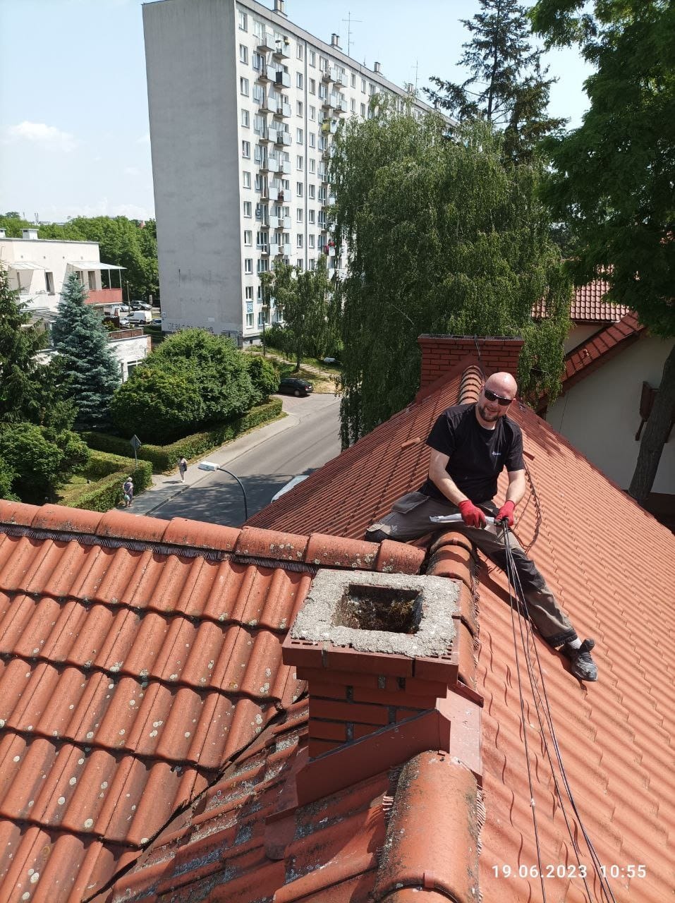 Pracownik w okularach na dachu z czerwonej dachówki, obok komin. W tle budynek mieszkalny i zieleń. Prace na wysokościach w słoneczny dzień.