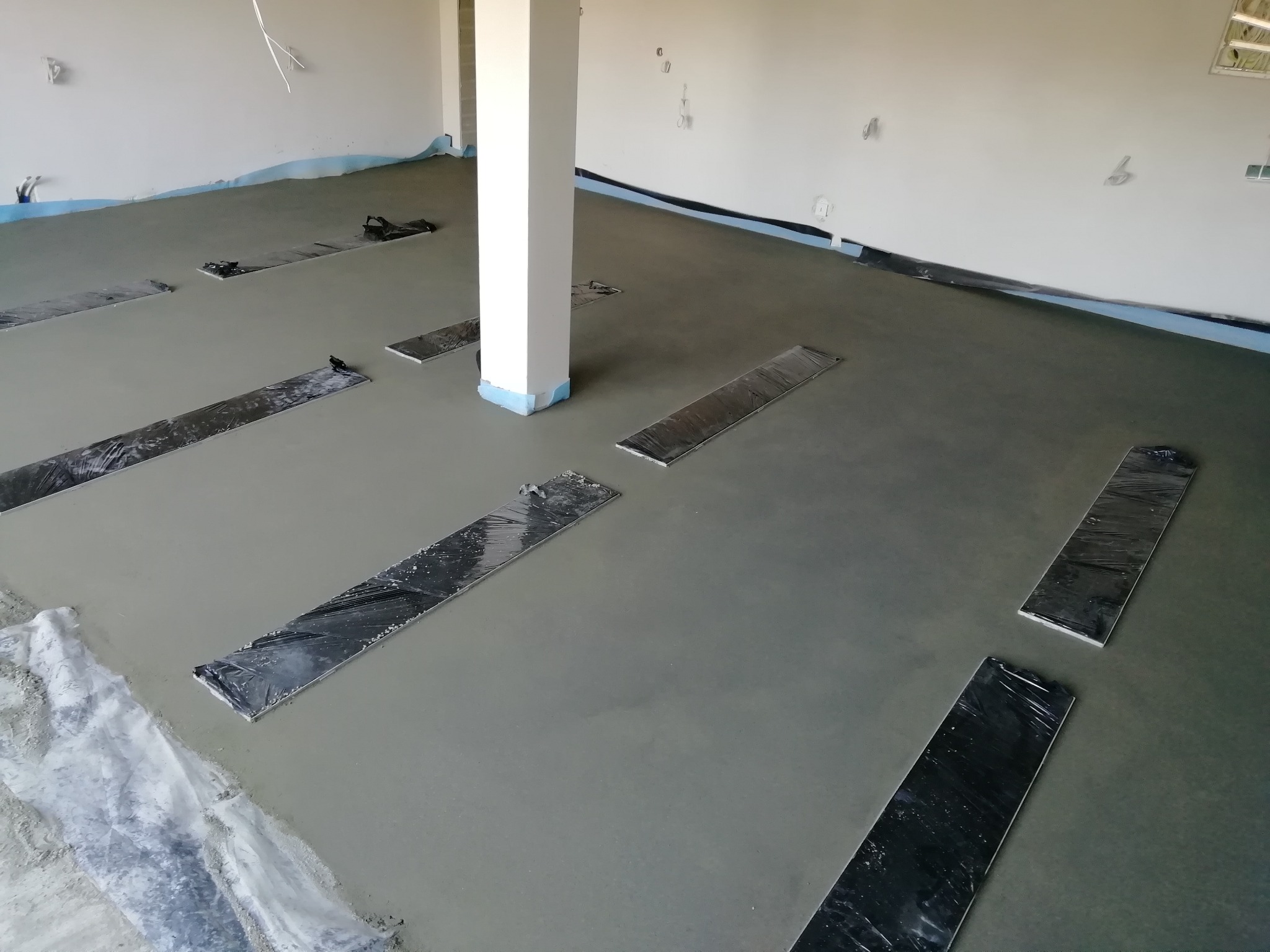 Świeżo wylana, jeszcze mokra posadzka jastrychowa z dylatacjami przy ścianach i wokół kolumny nośnej, zabezpieczonymi folią.
