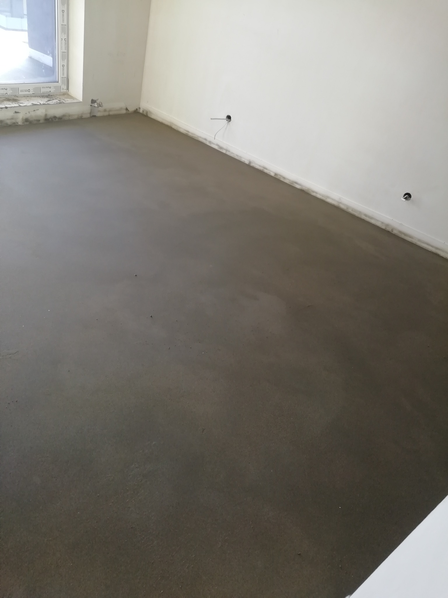 Świeżo wylana, mokra posadzka w jasnym pomieszczeniu z widocznymi otworami na kable w ścianie i folią okienną.