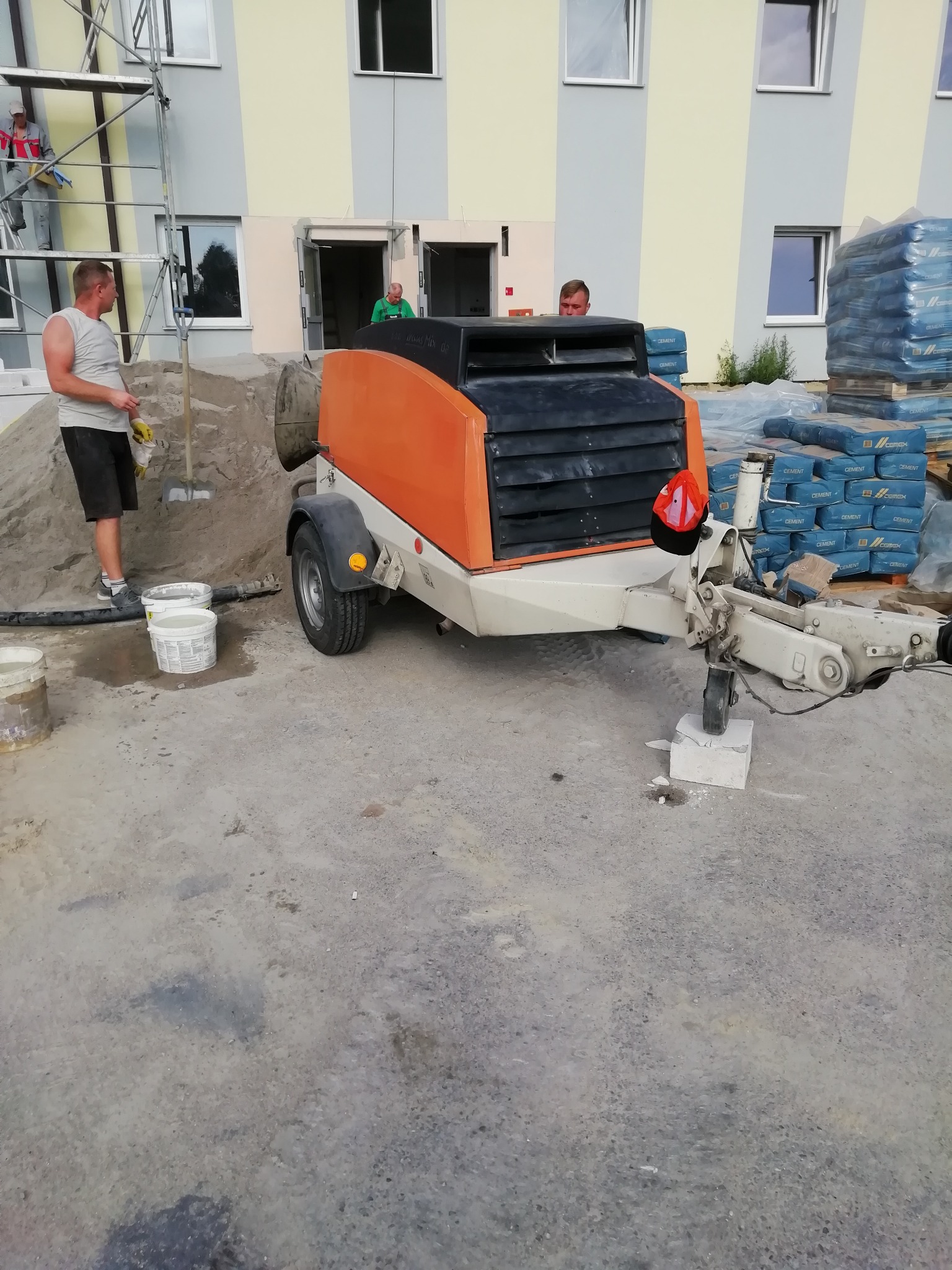 Pompa do wylewek z pomarańczową obudową, zaparkowana na placu budowy, obok sterty piasku i worków z cementem, pracownicy w tle.