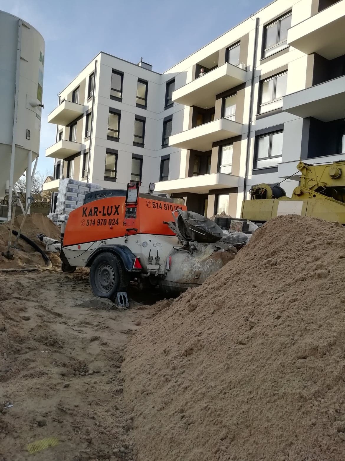 Pompa do wylewek z widocznym numerem telefonu firmy na tle nowoczesnego budynku mieszkalnego i hałdy piasku.