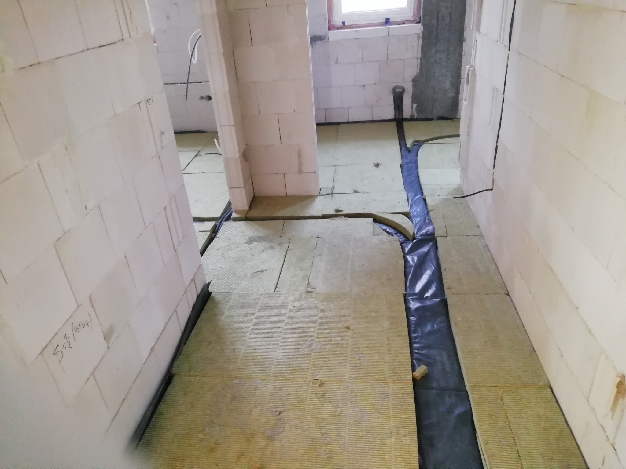 Wnętrze w trakcie prac: rozłożona warstwa izolacji na podłodze, zabezpieczona folią, widoczne ściany z bloczków i instalacje.