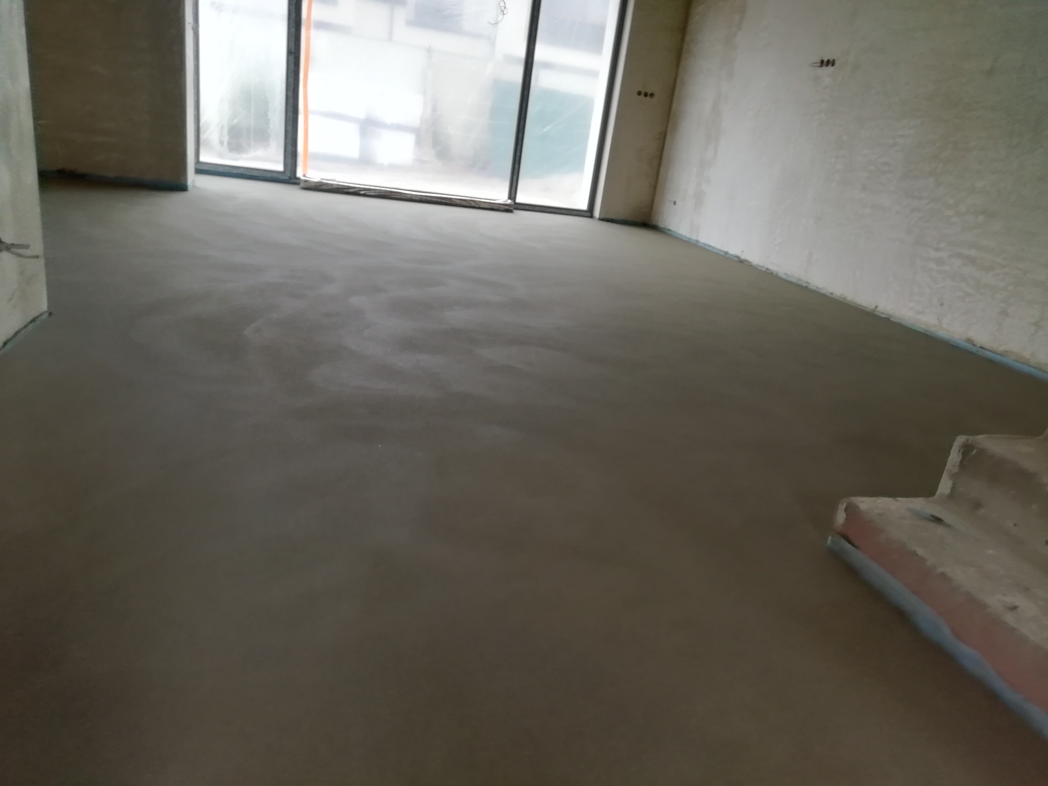 Świeżo wylana, gładka posadzka w surowym wnętrzu z dużym oknem, widoczne fragmenty schodów i ściany w stanie deweloperskim.
