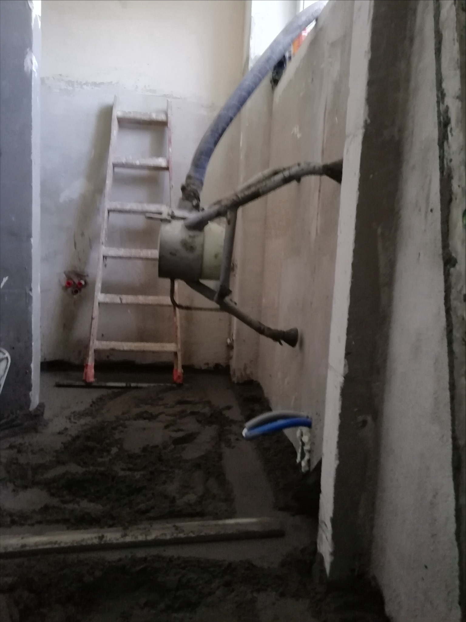 Aplikacja wylewki samopoziomującej w surowym wnętrzu, widoczna maszyna z wężami, drabina malarska oparta o ścianę, świeża wylewka na podłodze.