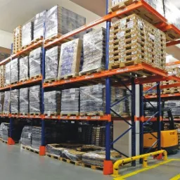 Pełne regały magazynowe z paletami i opakowanymi towarami, widoczny wózek widłowy i żółte bariery ochronne na szarej posadzce.