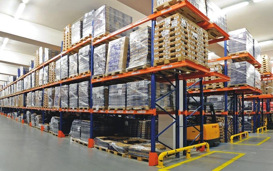 Pełne regały magazynowe z paletami i opakowanymi towarami, widoczny wózek widłowy i żółte bariery ochronne na szarej posadzce.