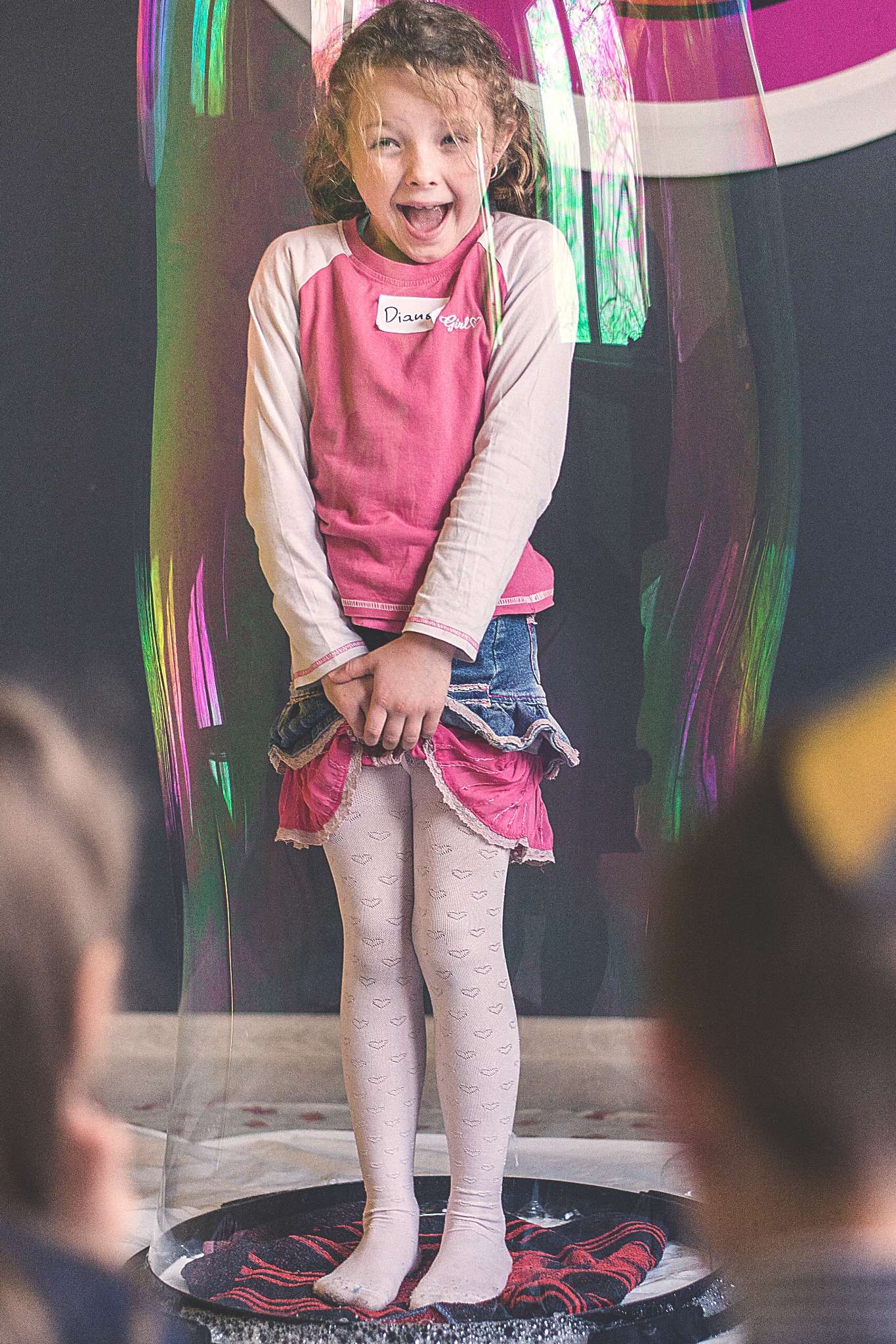 Uśmiechnięta dziewczynka z plakietką 'Diana Girl' w różowej bluzce i dżinsowej spódniczce, stojąca wewnątrz ogromnej, tęczowej bańki mydlanej na czarnej macie.