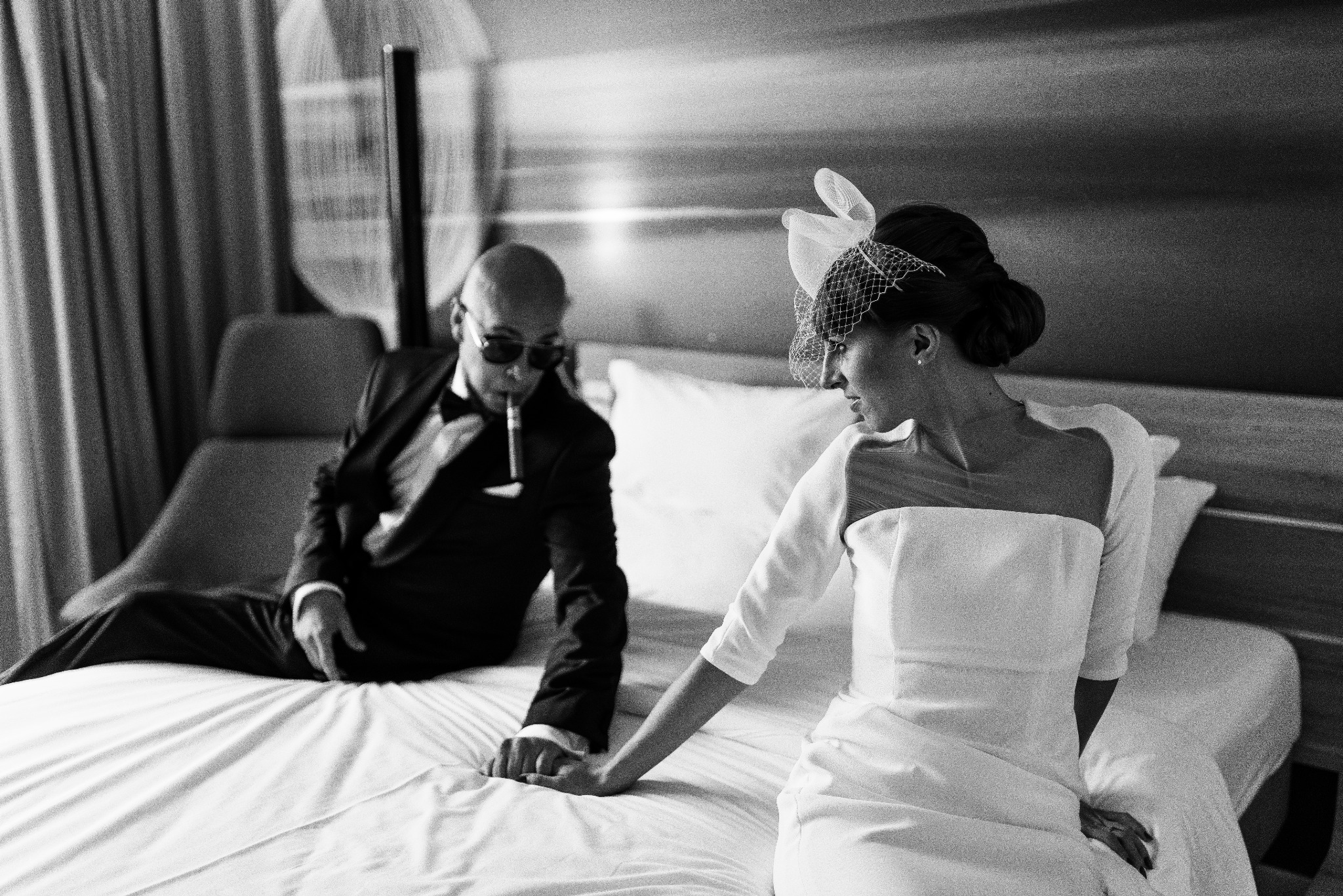 Czarno-białe zdjęcie pary młodej: panna młoda w sukni i woalce, pan młody w garniturze i okularach przeciwsłonecznych, trzymający fajkę, siedzą na łóżku trzymając się za ręce.