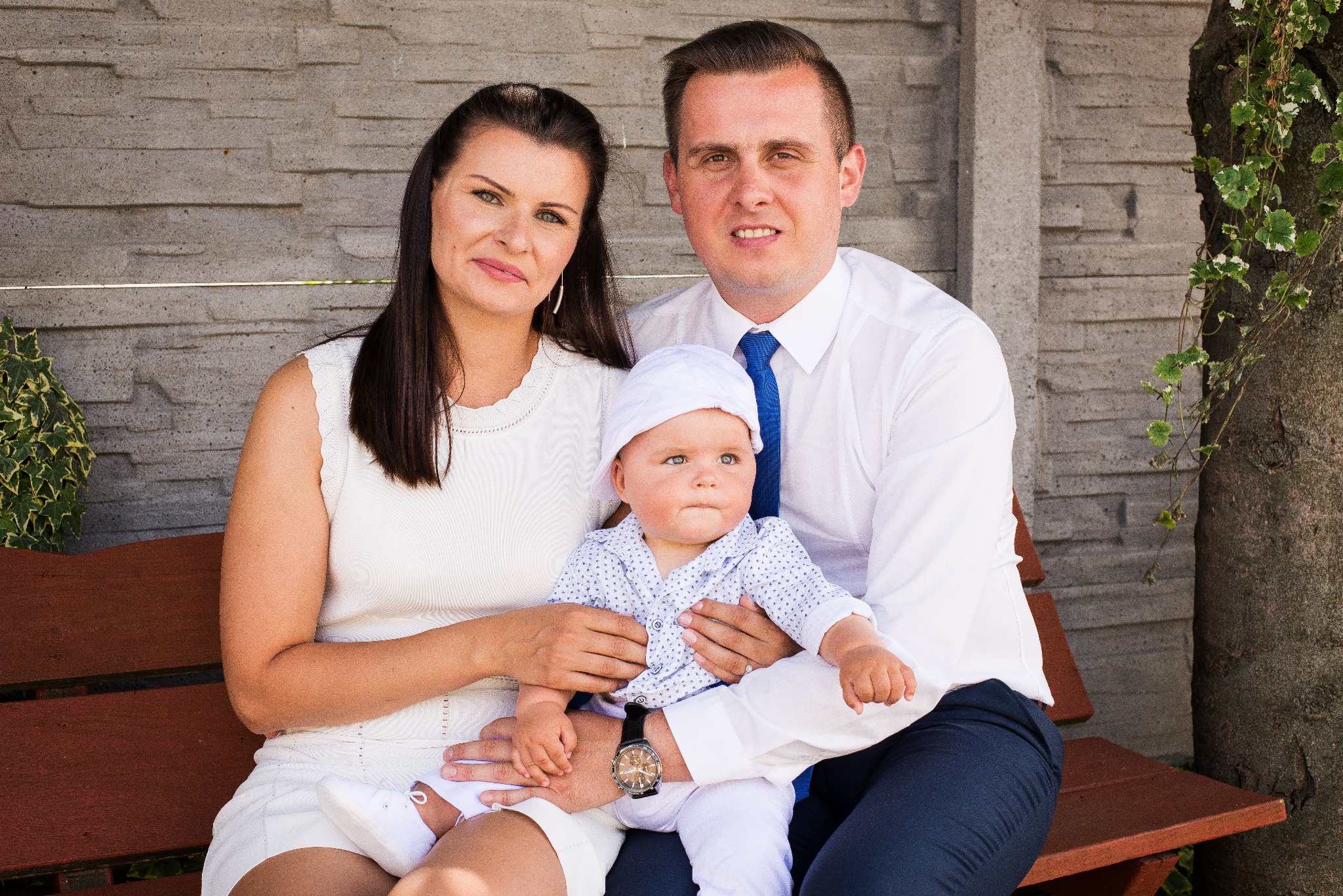 Rodzinne zdjęcie portretowe: mama, tata i dziecko w białych ubraniach siedzą na ławce na tle szarego muru i drzewa.