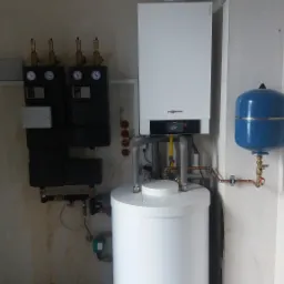 Nowoczesny, biały piec kondensacyjny Viessmann z podłączonym zasobnikiem ciepłej wody i niebieskim naczyniem wzbiorczym, zamontowany na szarej ścianie w kotłowni, obok czarnych paneli sterujących...