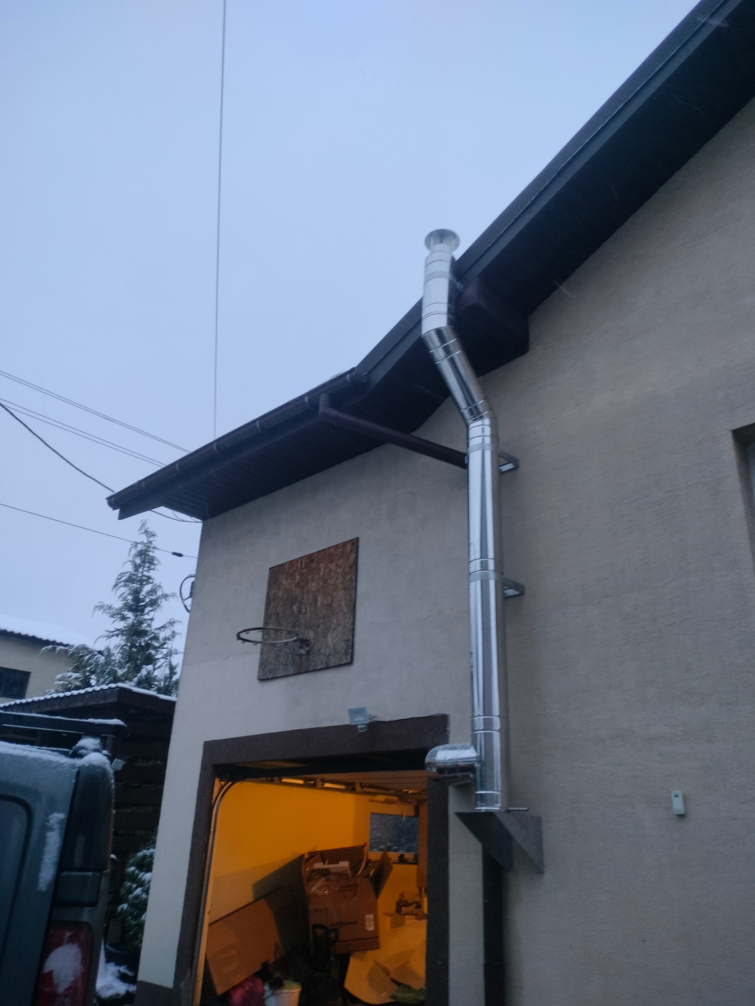 Zewnętrzny, pionowy, metalowy komin spalinowy, przymocowany do ściany budynku z garażem. Widoczne elementy montażowe i zakończenie komina. Pochmurne niebo w tle.