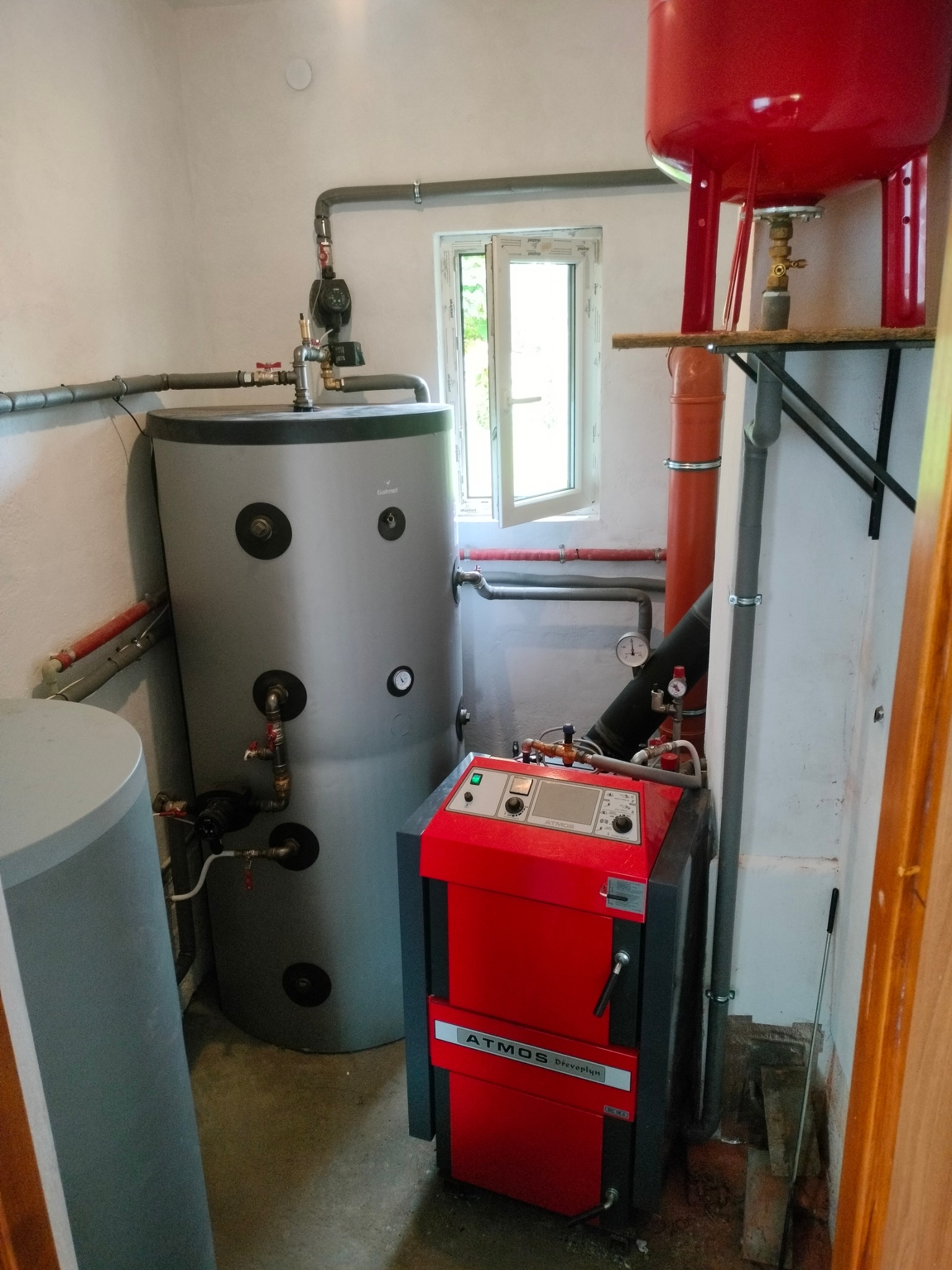 Zestaw kotła na paliwo stałe Atmos z zbiornikiem akumulacyjnym w kotłowni. Widoczne rury, zawory i manometry. Otwór okienny w tle.