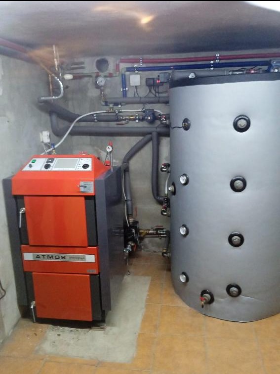 Pomarańczowo-szary piec Atmos obok srebrnego zbiornika akumulacyjnego w kotłowni. Widoczne rury i instalacje. Ujęcie w rogu pomieszczenia.