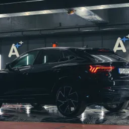 Błyszczący, czarny Audi Q3 Sportback po umyciu, widoczne odbicia w mokrej posadzce myjni, logo firmy detailingu na ścianie w tle.
