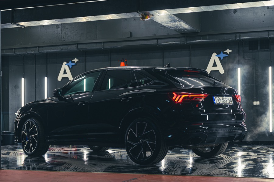 Błyszczący, czarny Audi Q3 Sportback po umyciu, widoczne odbicia w mokrej posadzce myjni, logo firmy detailingu na ścianie w tle.
