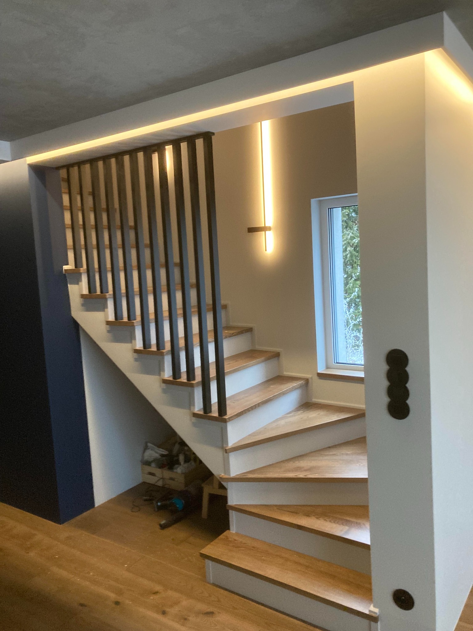 Nowoczesne schody z drewnianymi stopniami i czarną, minimalistyczną balustradą, oświetlone listwami LED w suficie i pionowym kinkietem, widok z dołu na zakręcające schody.