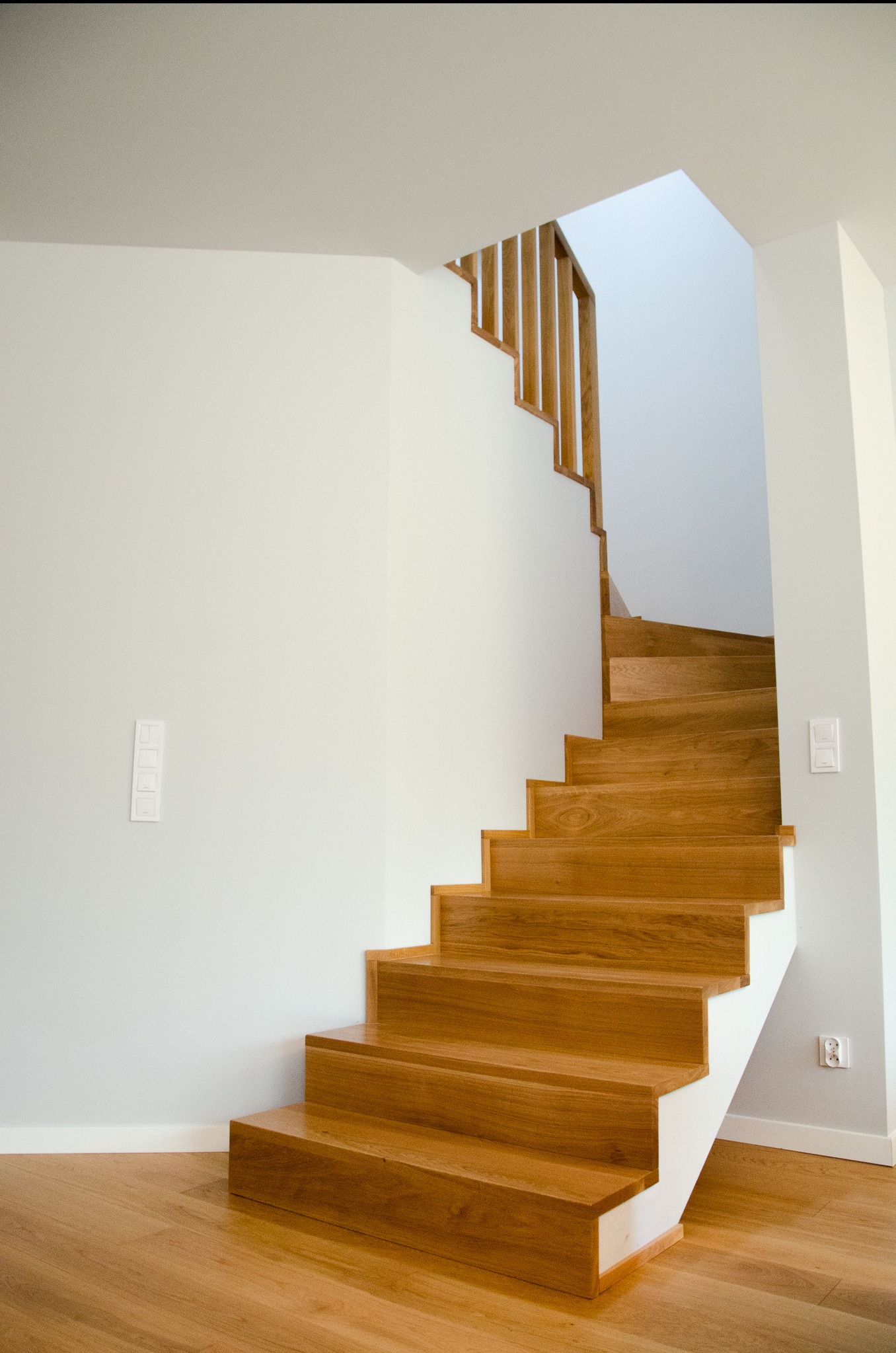 Nowoczesne, jasne wnętrze z drewnianymi schodami i ażurową balustradą, widok z dołu na białe ściany i drewnianą podłogę.