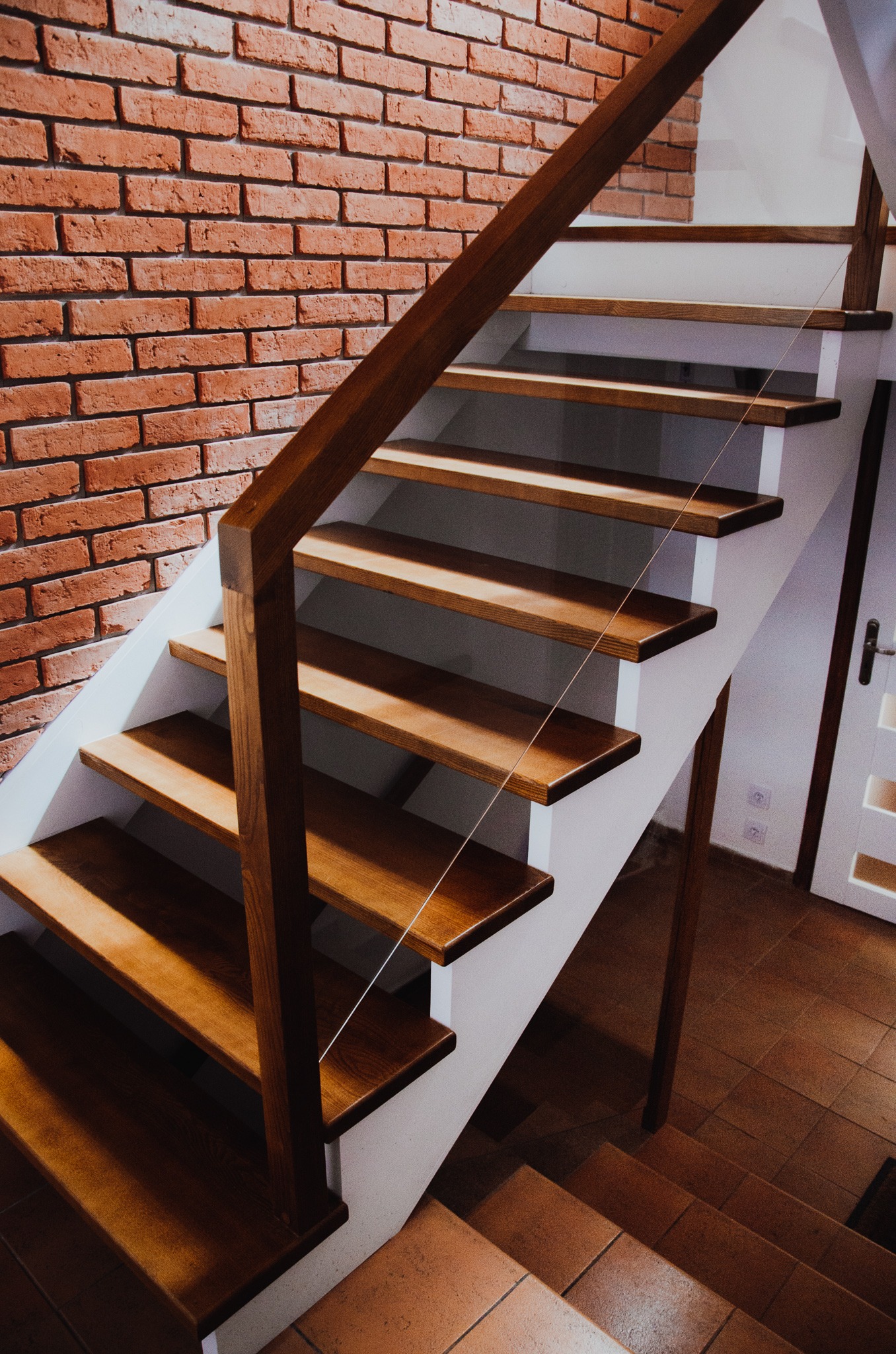 Nowoczesne schody z drewnianymi stopniami, białą konstrukcją i ceglaną ścianą w tle, oświetlone ciepłym światłem.