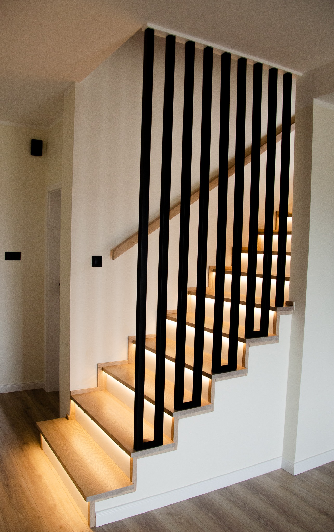 Nowoczesne schody z podświetleniem LED pod stopniami i czarną, pionową balustradą, widok z dołu.