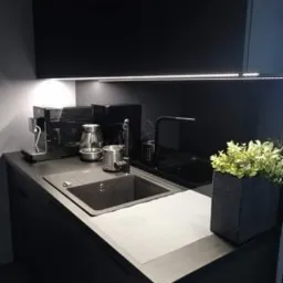 Nowoczesna, czarna kuchnia z oświetleniem LED podszafkowym, zlewem ze stali nierdzewnej i rośliną doniczkową w szarej osłonce, po sprzątaniu.