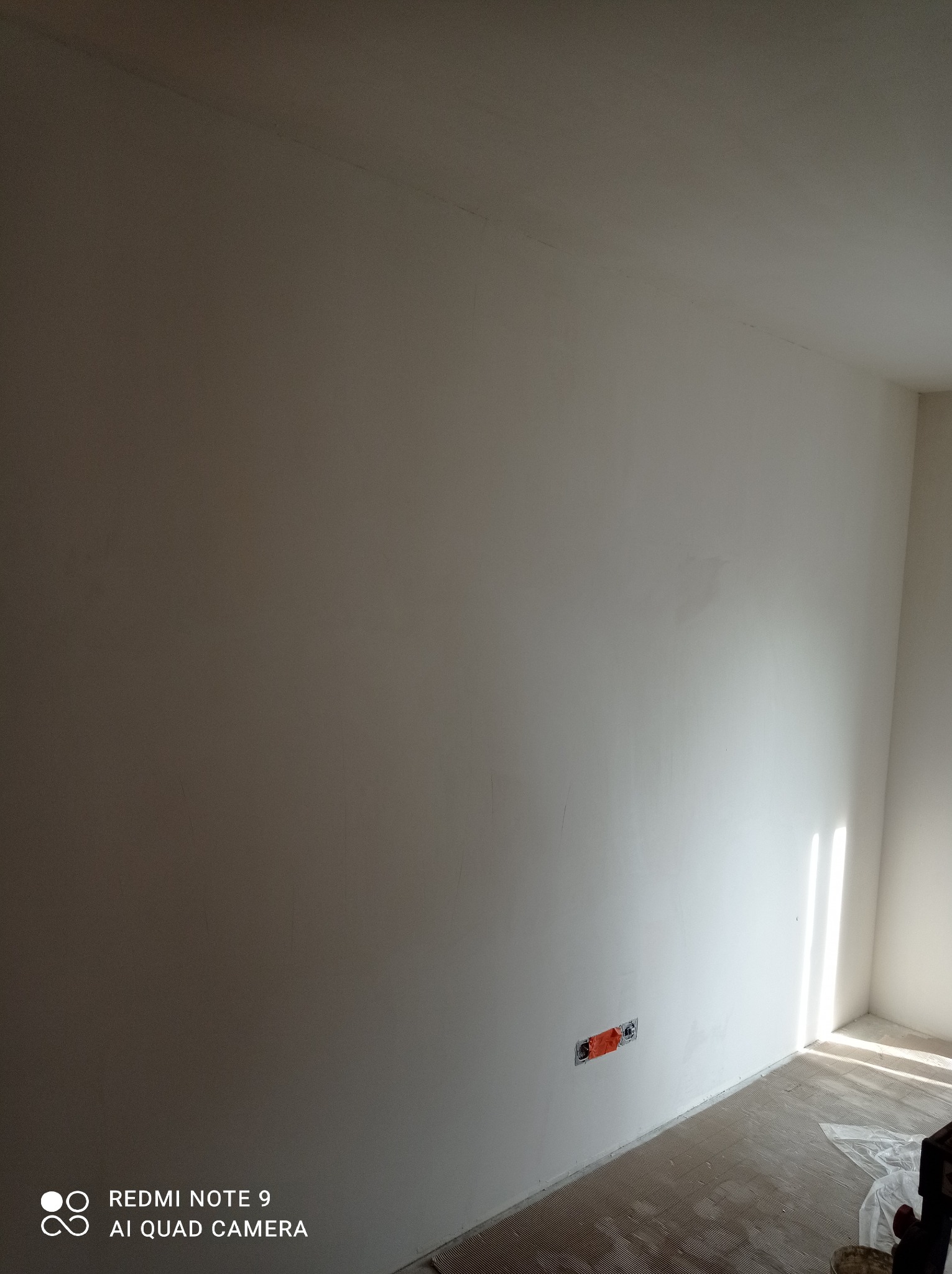Ściana po szpachlowaniu i malowaniu na biało z widoczną puszką elektryczną przed montażem osprzętu, zarys pod sufitem oraz nierówności na podłodze sugerują trwający remont.