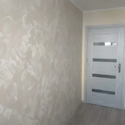 Ściana z dekoracyjnym efektem malowania w odcieniach beżu i bieli, widoczna szara wewnętrzna stolarka drzwiowa z poziomymi przeszkleniami i panele podłogowe w kolorze jasnego drewna.