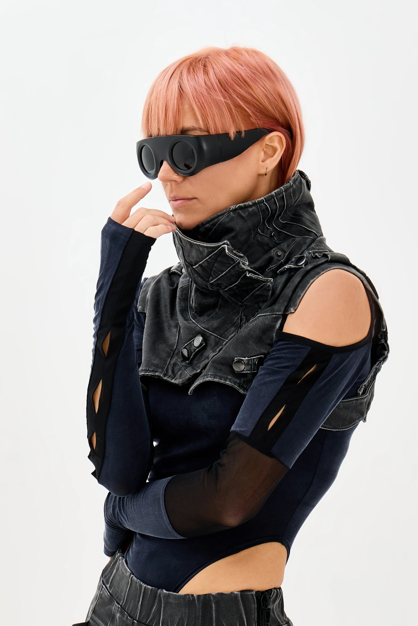 Kobieta z różowymi włosami w awangardowym stroju z czarną kamizelką z wysokim kołnierzem i futurystycznymi okularami, w zamyślonej pozie na białym tle.