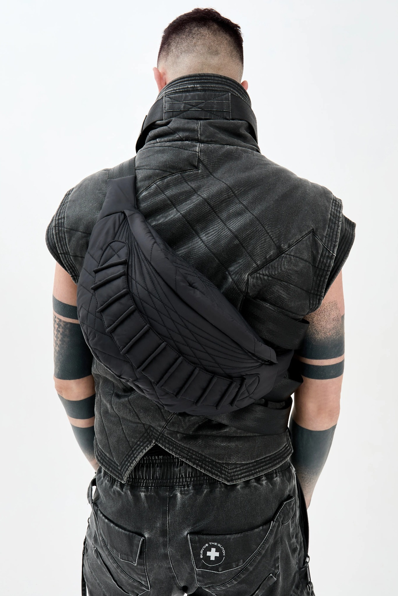 Mężczyzna tyłem w czarnym, postarzanym komplecie odzieży: kamizelka z pikowaniem i spodnie cargo z naszywką, z czarną, pikowaną torbą nerką na biodrach.