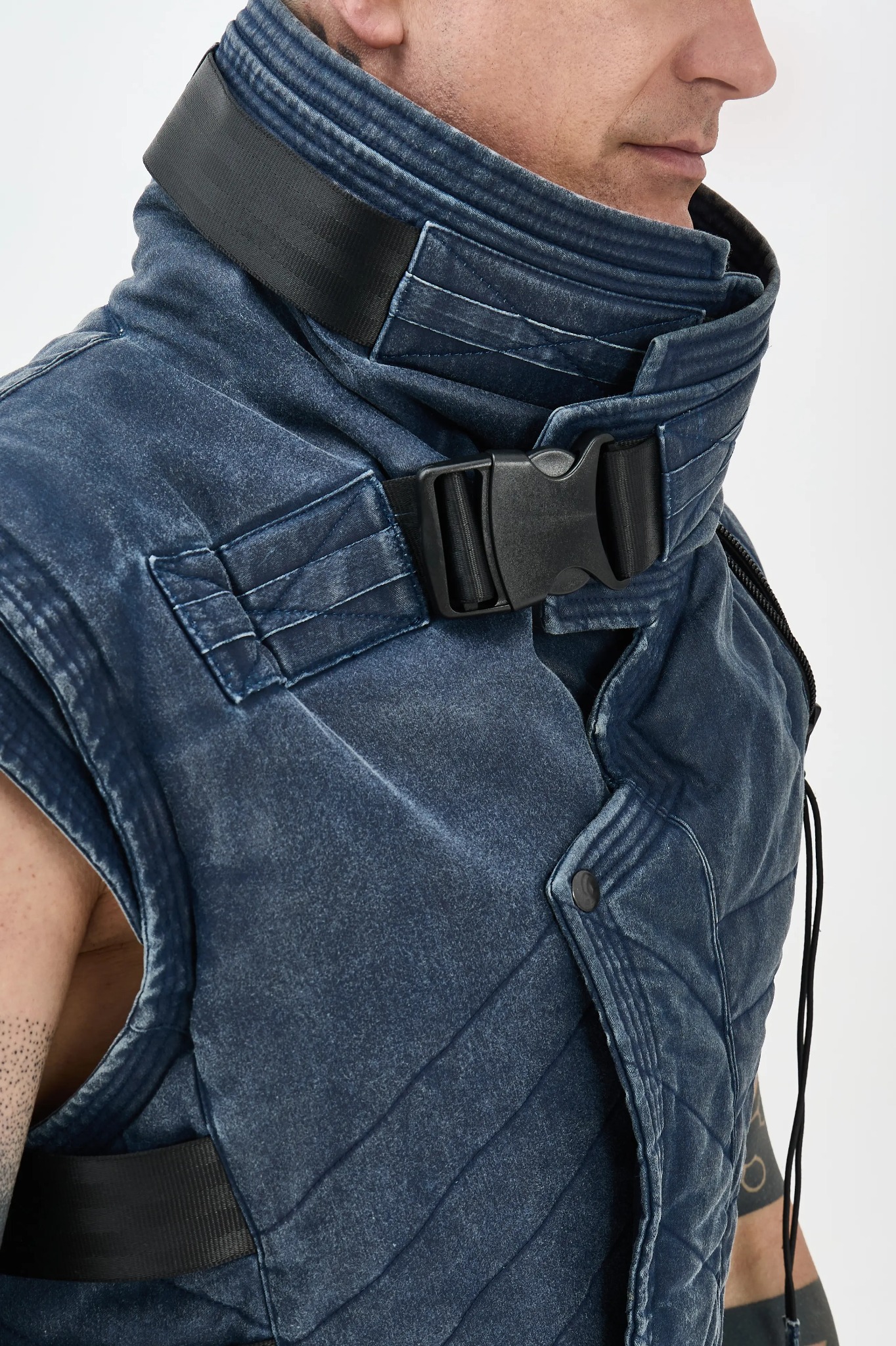 Mężczyzna w denimowej, pikowanej kamizelce z wysokim kołnierzem zapinanym na klamrę, widok z boku, fragment tatuażu na ramieniu.