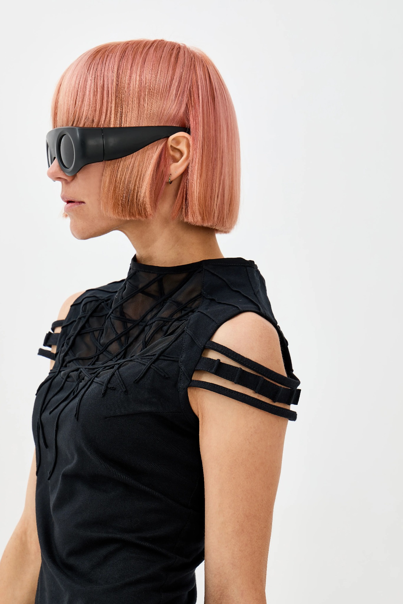 Kobieta z różowymi włosami w czarnej, awangardowej bluzce z siatki i futurystycznych okularach, profil, na białym tle.