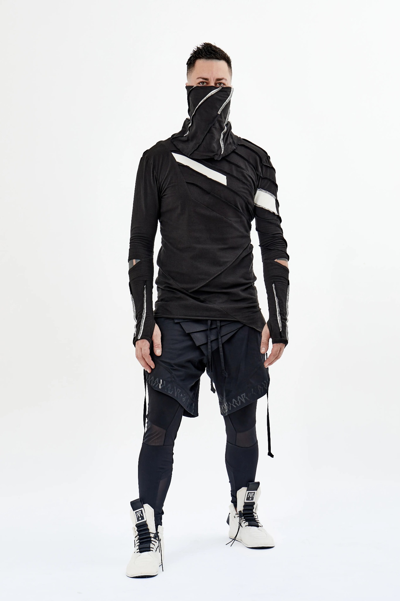 Mężczyzna w awangardowym, czarnym stroju z zakrytą twarzą, prezentujący unikalny design ubrania z asymetrycznymi elementami i nietypowymi wykończeniami na jasnym tle.