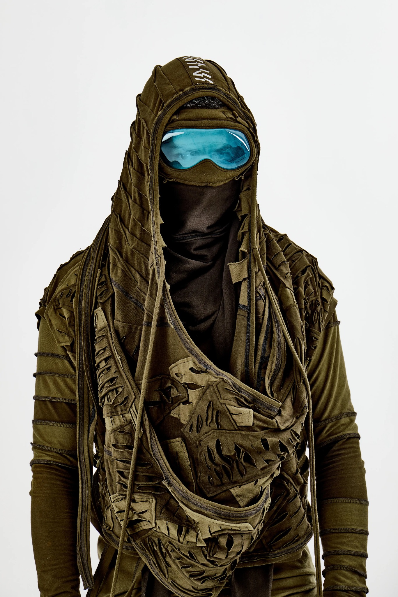 Awangardowy strój w odcieniach zieleni z kapturem, ozdobnymi wycięciami i błękitnymi goglami, na białym tle.