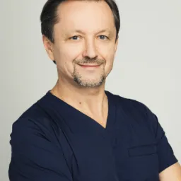 Portret uśmiechniętego technika dentystycznego w granatowym uniformie, z rękami skrzyżowanymi na piersi, na neutralnym tle.